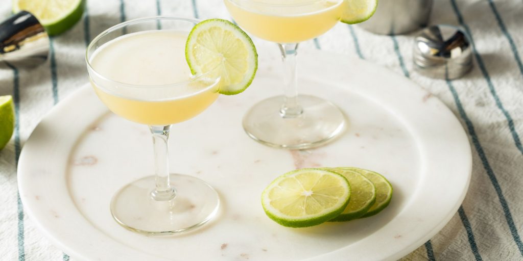 Close up top view of two Gimlet Cocktails in coupe glasses, garnished with lime wheels, and presented on a white platter set on a striped blue and white tablecloth