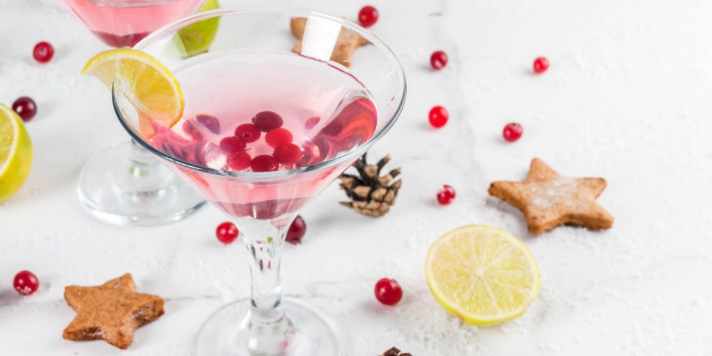 A pretty Cranberry Martini garnished with fresh cranberries, against a white backdrop studded with pine cones and ginger biscuits in the shape of stars