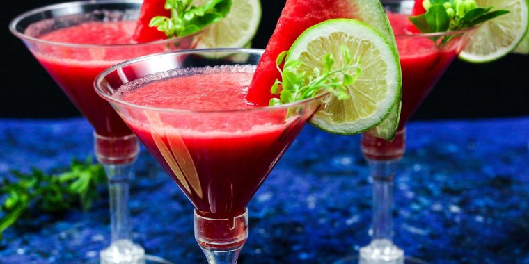 Two Watermelon Martinis garnished with a lemon wedge and slice of watermelon each, against a blue backdrop