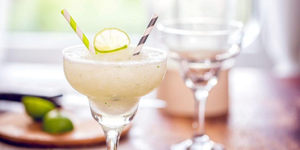 Close up of a Frozen Lime Margarita with a striped paper straw, with a second, empty glass in the background next to a cutting board with cut limes