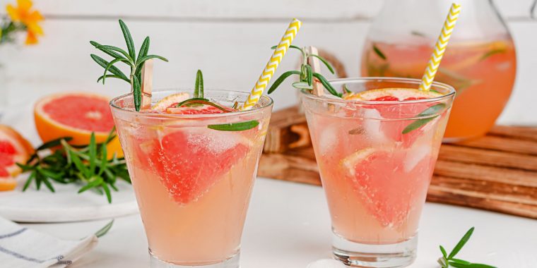 Close up of a pair of Greyhound Cocktails garnished with fresh grapefruit, on a white surface, with sliced grapefruit and a jug of juice in the background