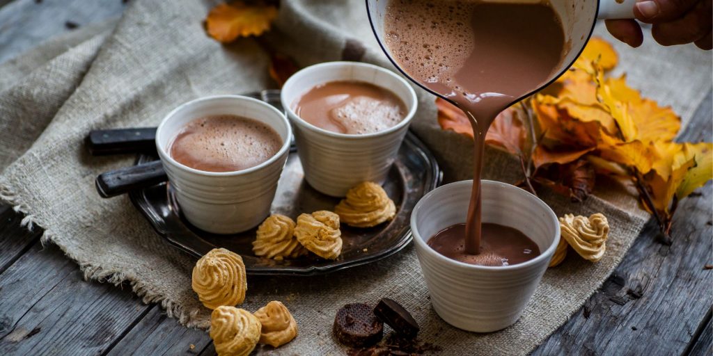 Top view of three mugs of Ancho Reyes Hot Chocolate being poured. presented on a rustic wooden surface partially covered in a raw linen table cloth, with small cookies scattered around
