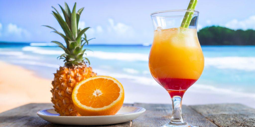 Close-up of a Malibu Sunset cocktail in a Hurricane glass with a view of the ocean in the background and a plate with a sliced orange and fresh pineapple to the left