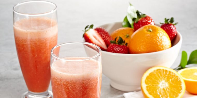 Close-up of a refreshing pair of Strawberry Mimosa cocktails against a white backdrop with a bowl of oranges and strawberries in the frame