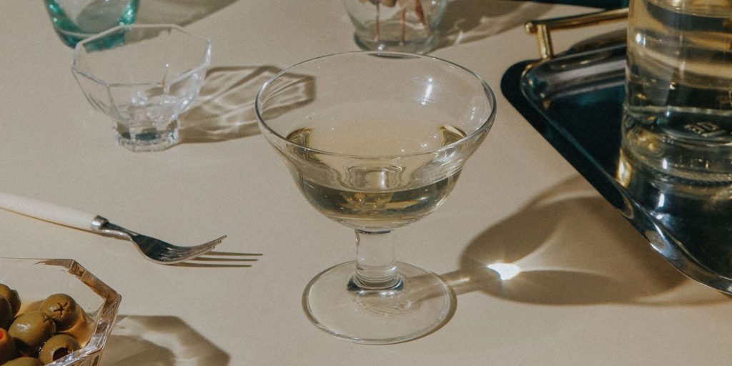 Close-up of stylish and simple Irish Martini cocktail on a white tablecloth, surrounded by assorted vintage cultery and crockery, including a bowl of green olives
