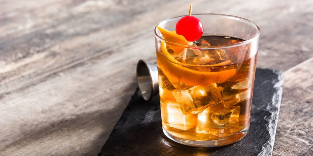 Close up of an Irish Old Fashioned Cocktail garnished with a cherry and an orange twist, set against a slate grey backdrop