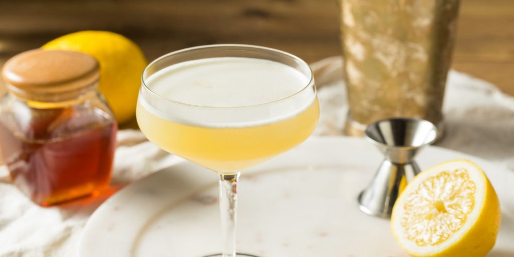 Close-up of a zingy Pot O' Gold cocktail on a white plate on a wooden surface, surrounded by a jar of honey, a cut lemon and a metal cocktail jigger