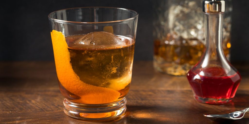 Vieux Carré Cocktail presented in a rocks glass over a large, circular piece of ice, garnished with a citrus peel, and placed on a wooden surface