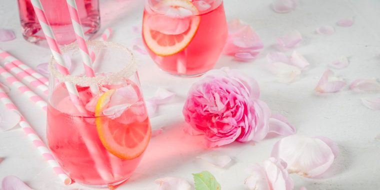 Three pretty pink Mother's Day cocktails against a white backdrop with a pink rose and some petals scattered for a touch of whimsy