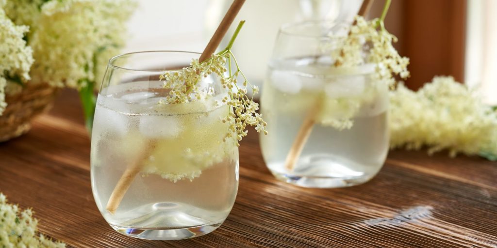 Two Elderflower & Herb Cooler cocktails with bamboo straws, garnished with extra Elderflower blooms against a wooden backdrop