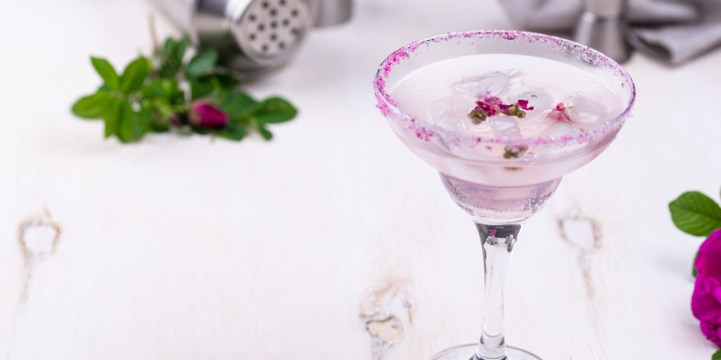 A pretty Honey Rose Margarita cocktail with a sparkly rim, garnished with rose petals against a white backdrop with some roses to the side