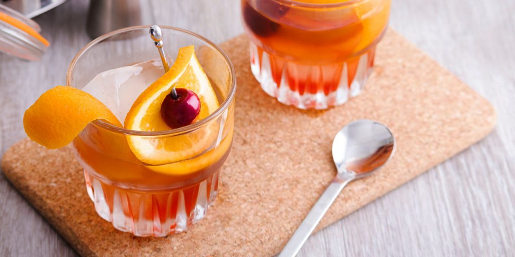 Two Marzipan Old Fashioned cocktails on a cork surface, garnished with an orange twist and cherry each