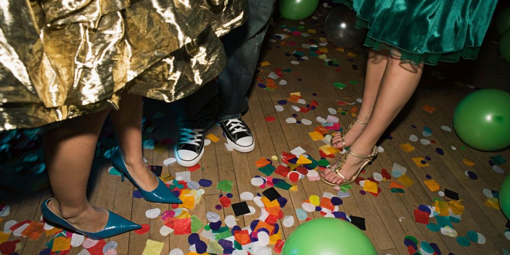 The feet of a group of friends gathered on a confetti-strewn floor at an Oscar viewing party wearing party dresses and colorful shoes