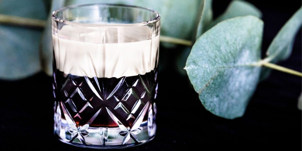 A creamy Baby Guinness Shot against a dark backdrop decorated with a sprig of pennygum