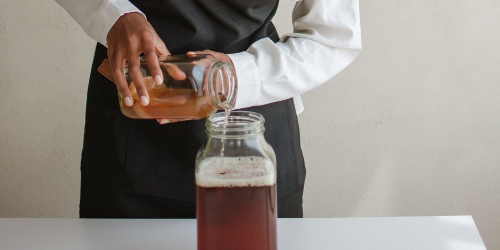 A front view of a woman in a white shirt and black apron measuring liquid from a smaller container into a large container