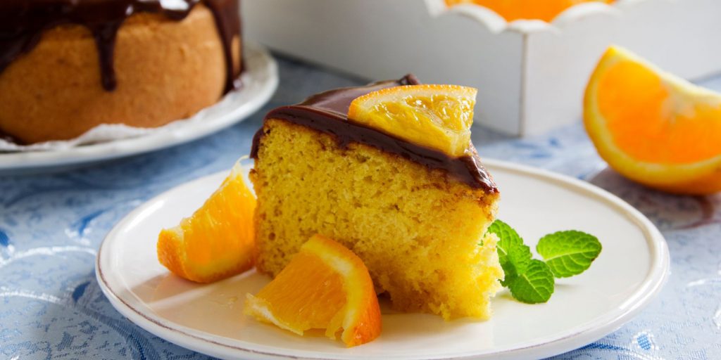 Close up of a slice of chocolate orange cake garnished with fresh slices of orange on a white plate with the rest of the cake and orange wedges in the background