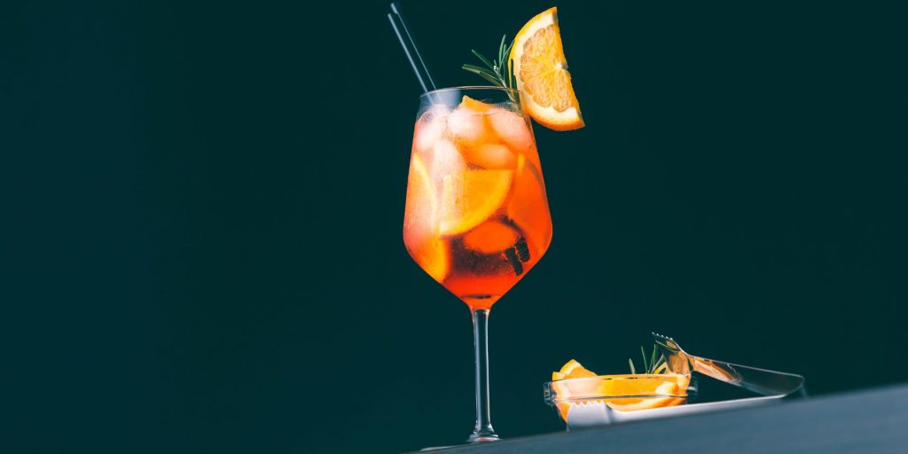 Mid-range shot of an orange cocktail in a wine glass garnished with a fresh slice of orange against a dark background