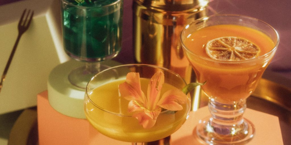 A close up of two retro-looking orange cocktails on a dusty pink podium with a golden cocktail shaker in the background 