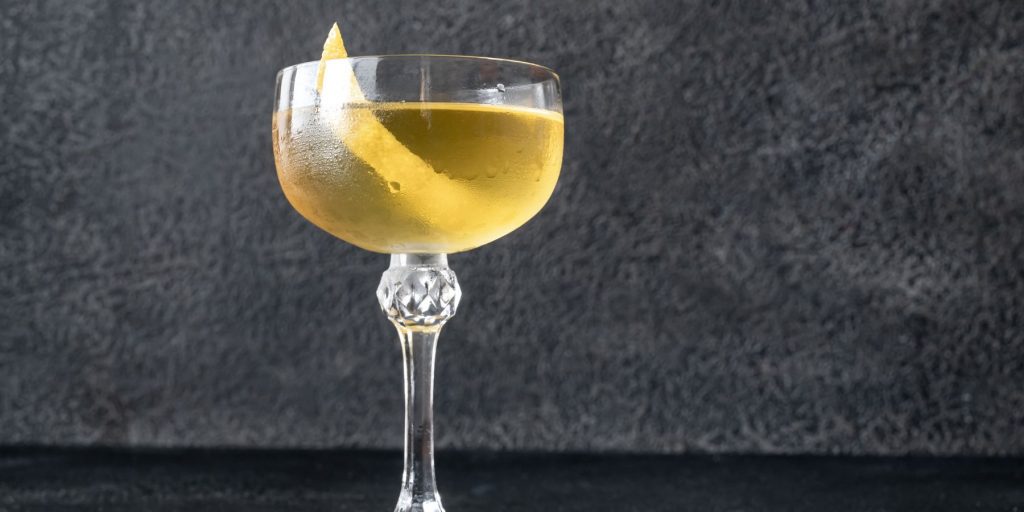 Close-up view of a yellow-tinged Alaska cocktail garnished with a lemon twist against a dark grey backdrop 