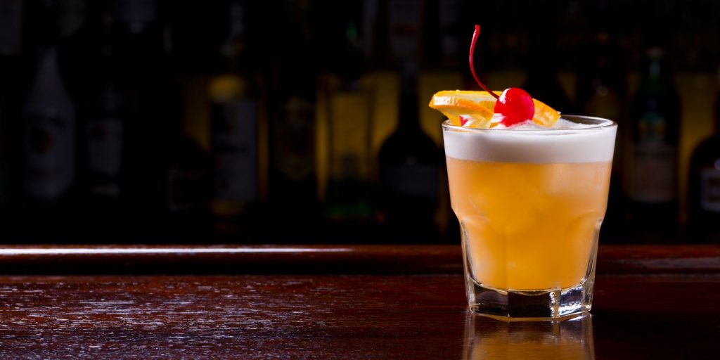 Front view of a Fernet Sour Cocktail garnished with a cherry on a wooden surface against a dark background
