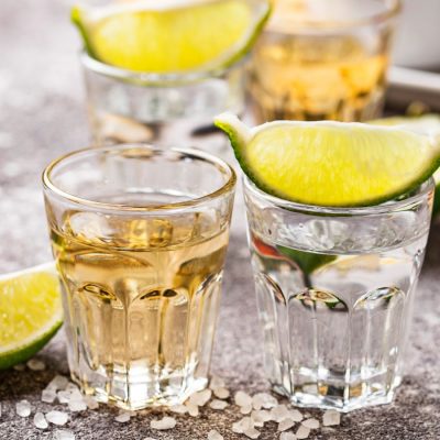 Close up of two shot glasses of tequla garnished with lemon wedges on a light grey background