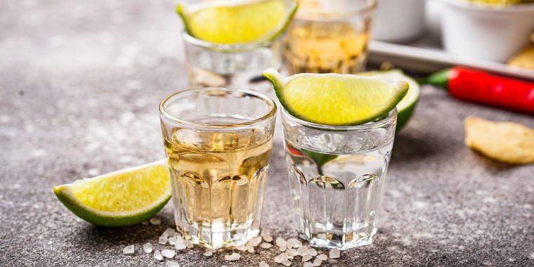 Close up of two shot glasses of tequla garnished with lemon wedges on a light grey background