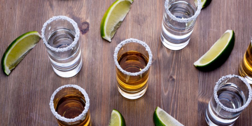 Top view of five shot glasses of tequila on a wooden surface interspersed with lime wedges