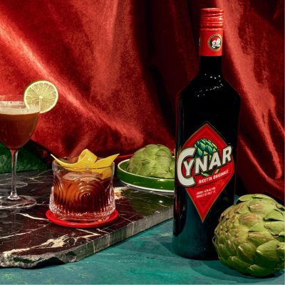 A sumptuous collection of retro lush cocktails posed next to a bottle of Cynar liqueur agains a blue marble backdrop draped with red velvet