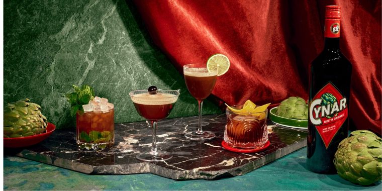 A sumptuous collection of retro lush cocktails posed next to a bottle of Cynar liqueur agains a blue marble backdrop draped with red velvet