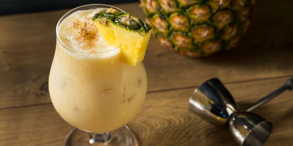 Close up of a Painkiller cocktail in a Hurricane glass garnished with a slice of pineapple on a wooden surface with a fresh pineapple towards the back and a metal jigger to the side