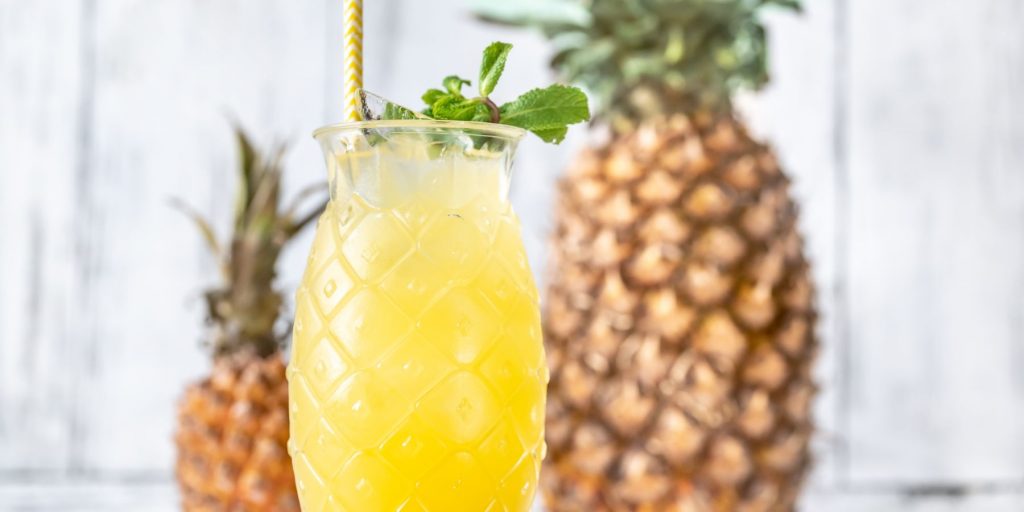 Close up of a Island Oasis cocktail in a glass shaped like a pineapple with two fresh pineapples in the background against a fresh white backdrop