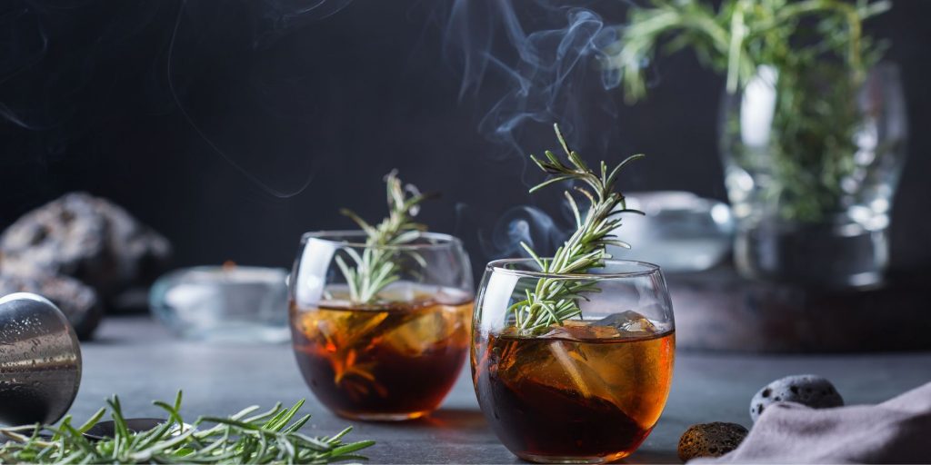 Front view of two Campfire Sours garnished with smoking rosemary against a dark grey backdrop with a batch of fresh rosemary in a vase 