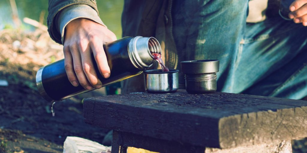 A close up of a hiker pouring himself a Mountain Daiquiri from a flask, on a wooden surface in an outdoor environment 