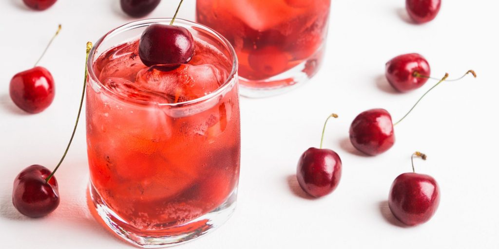 A pair of pretty Dirty Shirley cocktails in rocks glasses, garnished with fresh cherries, placed on a white surface
