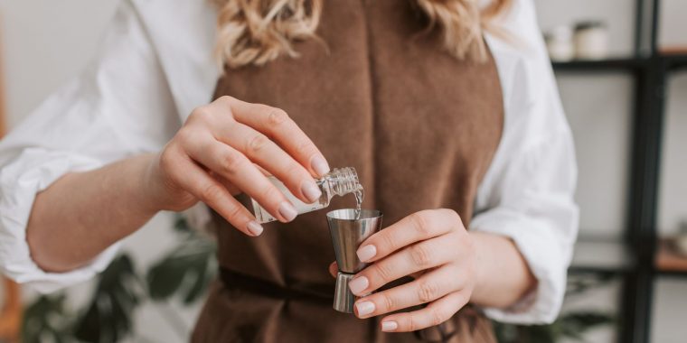 Close up front shot of a woman in a blonde woman in a brown apron pouring clear liquid into a metal jigger from a small bottle