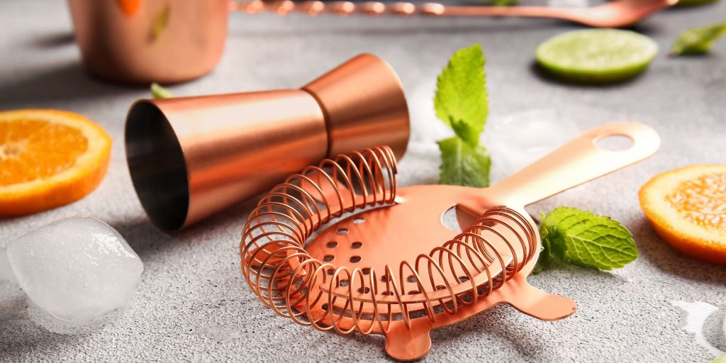 Close up front view of a copper jigger and cocktail strainer laid out on a white marble surface