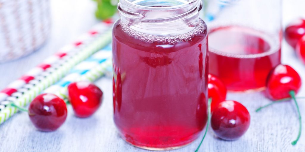 Close-up of a jar of cherry juice on a white surface surrounded by fresh cherries