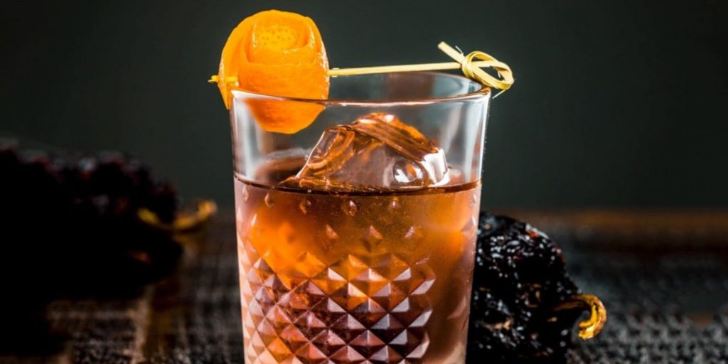 Close up of an Ancho Reyes Old Fashioned Cocktail in a rocks glass, garnished with an orange twist on a bamboo cocktail pick