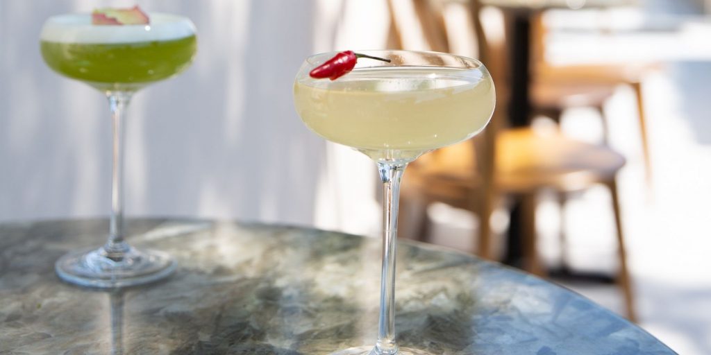 Close up of a zingy Bee Sting cocktail garished with a red chili on an outdoor table in the bright sunlight along with a green cocktail in the background