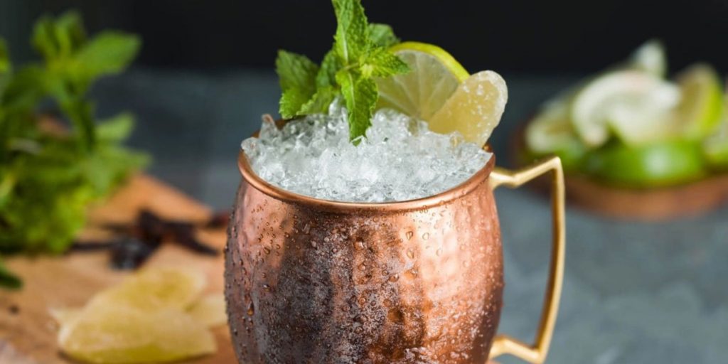 Close up of a Spicy Mexican Mule cocktail in a copper mug against a grey backdrop, garnished with fresh mint and lemon slices