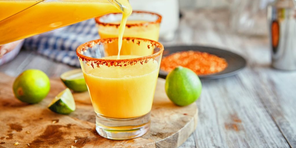 Close up of two glasses of Spicy Pineapple Margarita on a wooden table top surrounded by fresh limes and rimmed in tajin