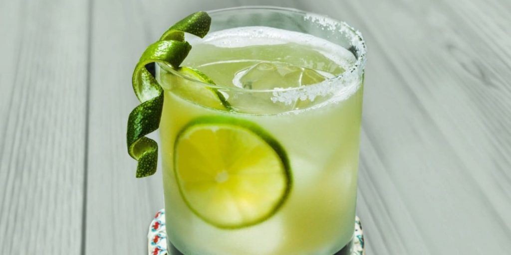 Close up of a Spicy Verde Margarita garnished with a cucumber twist against a light wood backdrop