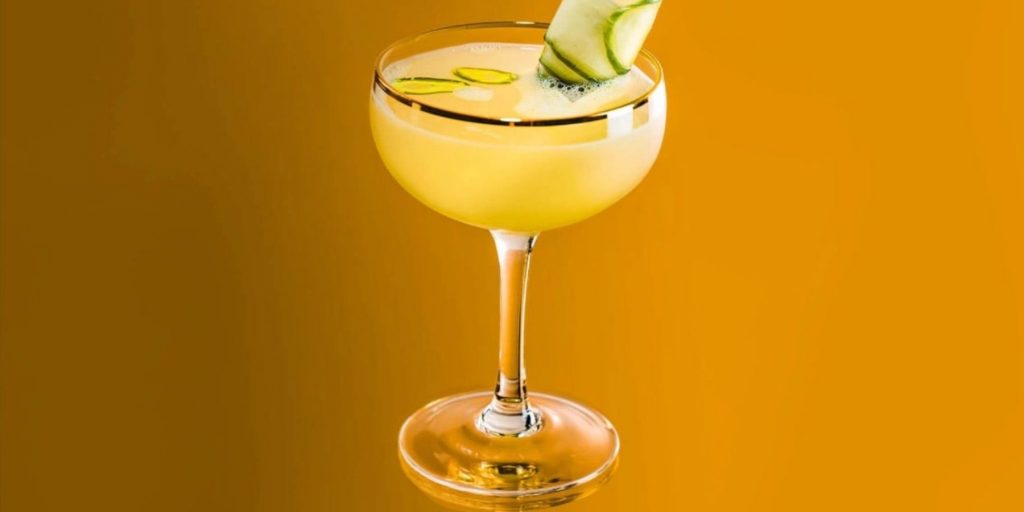 Close up of a Verde Gimlet in a coupe glass with a gold rim against a deep yellow backdrop