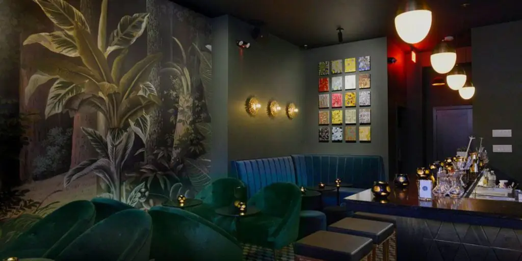 Moody shot of the interior of the Sugar Monk cocktail bar in NYC featuring lovely tropical wallpaper and plush seating