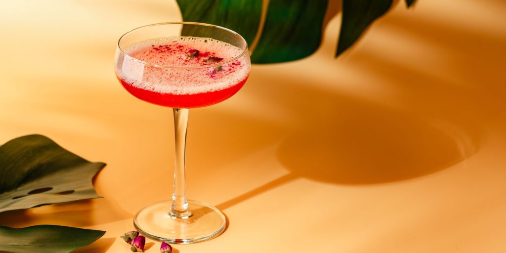Raspberry Gimlet in a coupe glass against a yellow backdrop with foilage embellisments as decoration