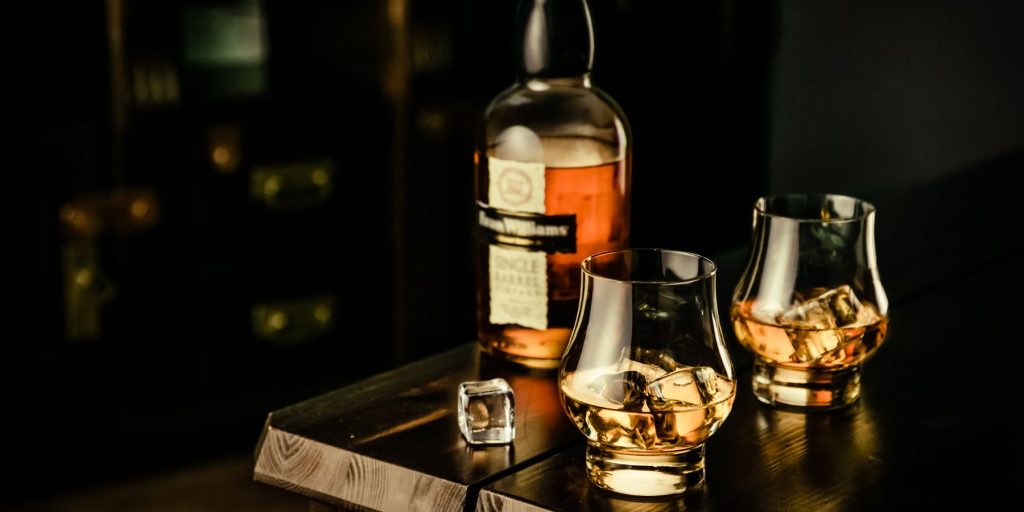 Two shifter whiskey glasses next to a bottle of whiskey on a table in a pub environment against a black backdrop