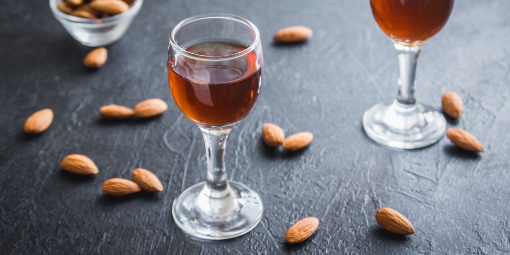 Two small snifters of amaretto on a dark grey background surrounded by a scaterring of fresh almonds