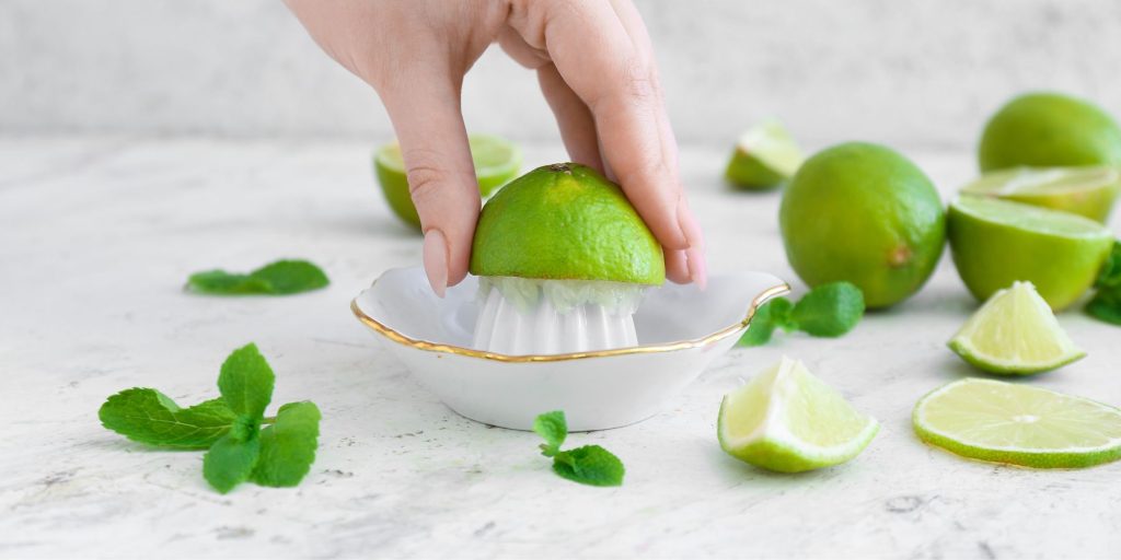Close up of a woman's hand squeezing fresh limes using a dainty ceramic juicing tray on a white backdrop