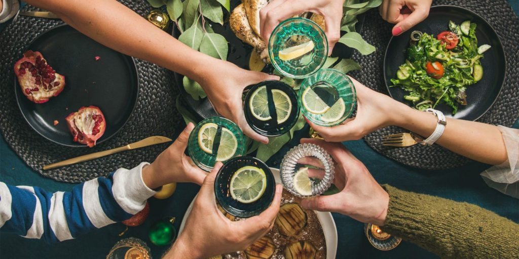 Top view of a group of friends clinking together festive Christmas cocktails over a table filled with holiday foods