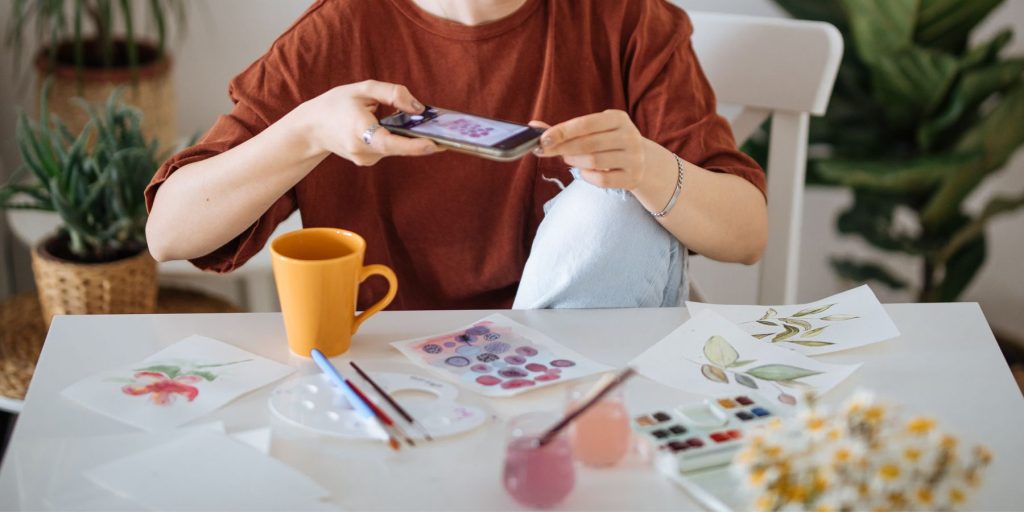 Close-up image of a woman taking a digital snap of a drawing collage she is busy making in a light brigh home lounge environment